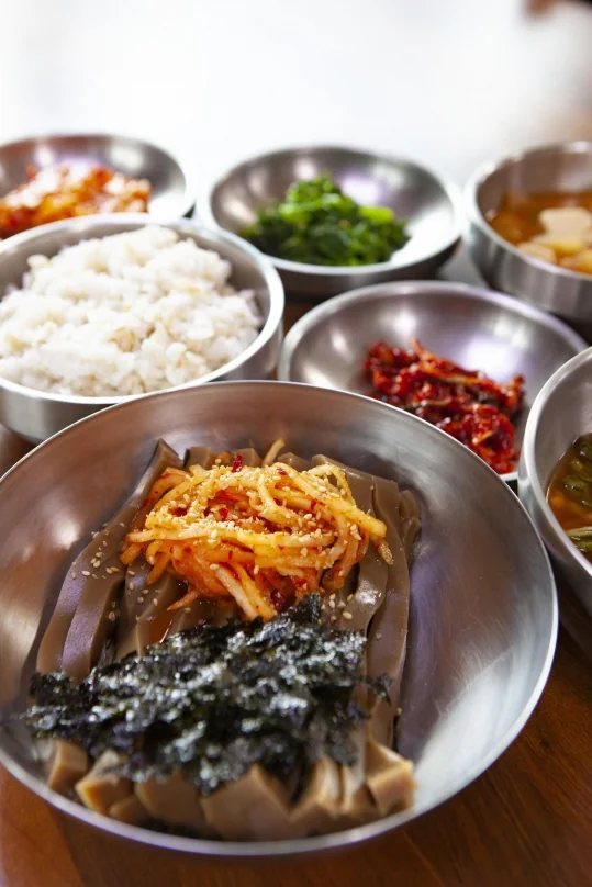 도토리묵밥 사발 — 붉은 무침 고명과 김가루, 뒤로 보리밥과 반찬